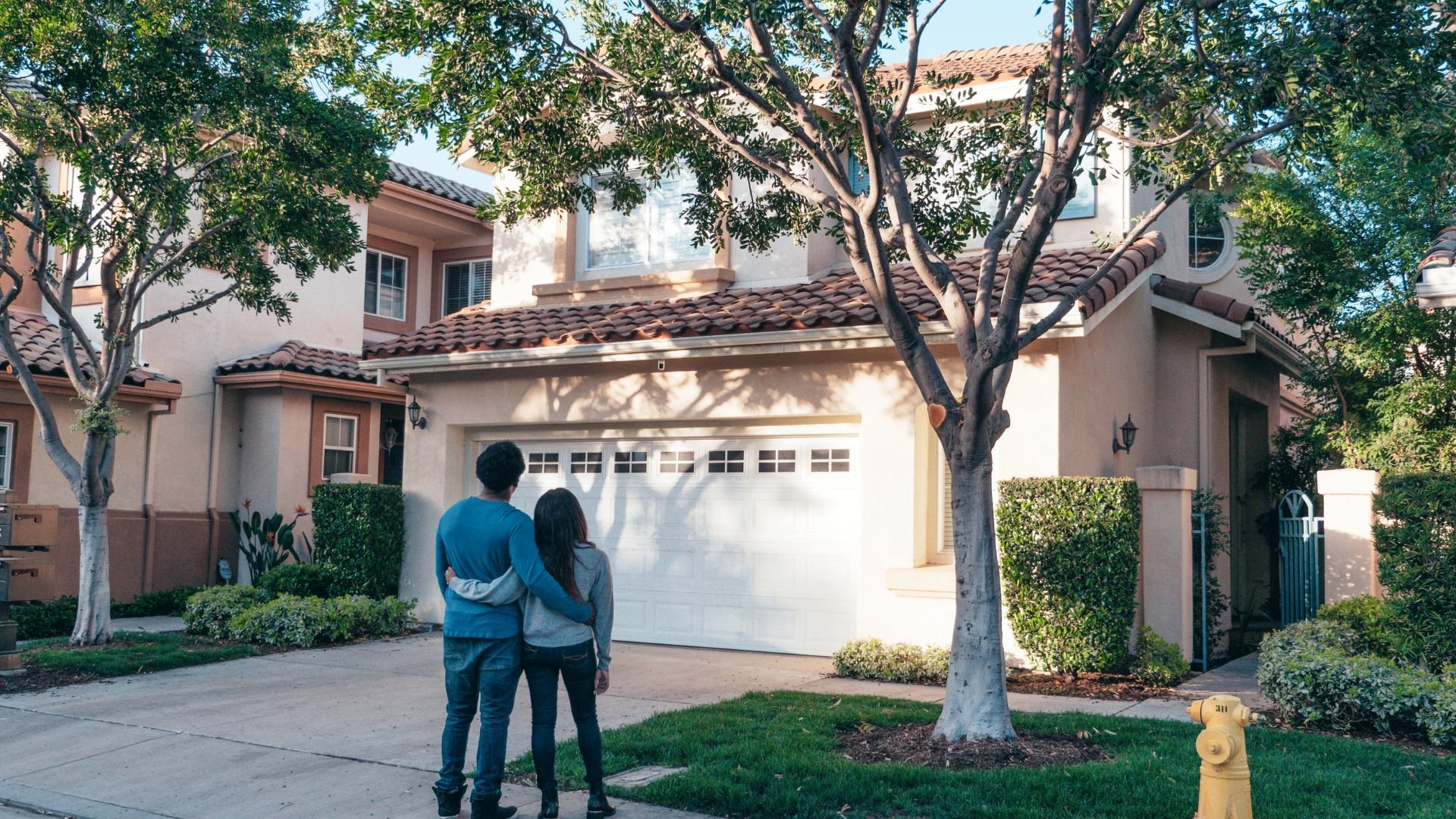 Two people standing in front of suburban house with trees and white garage